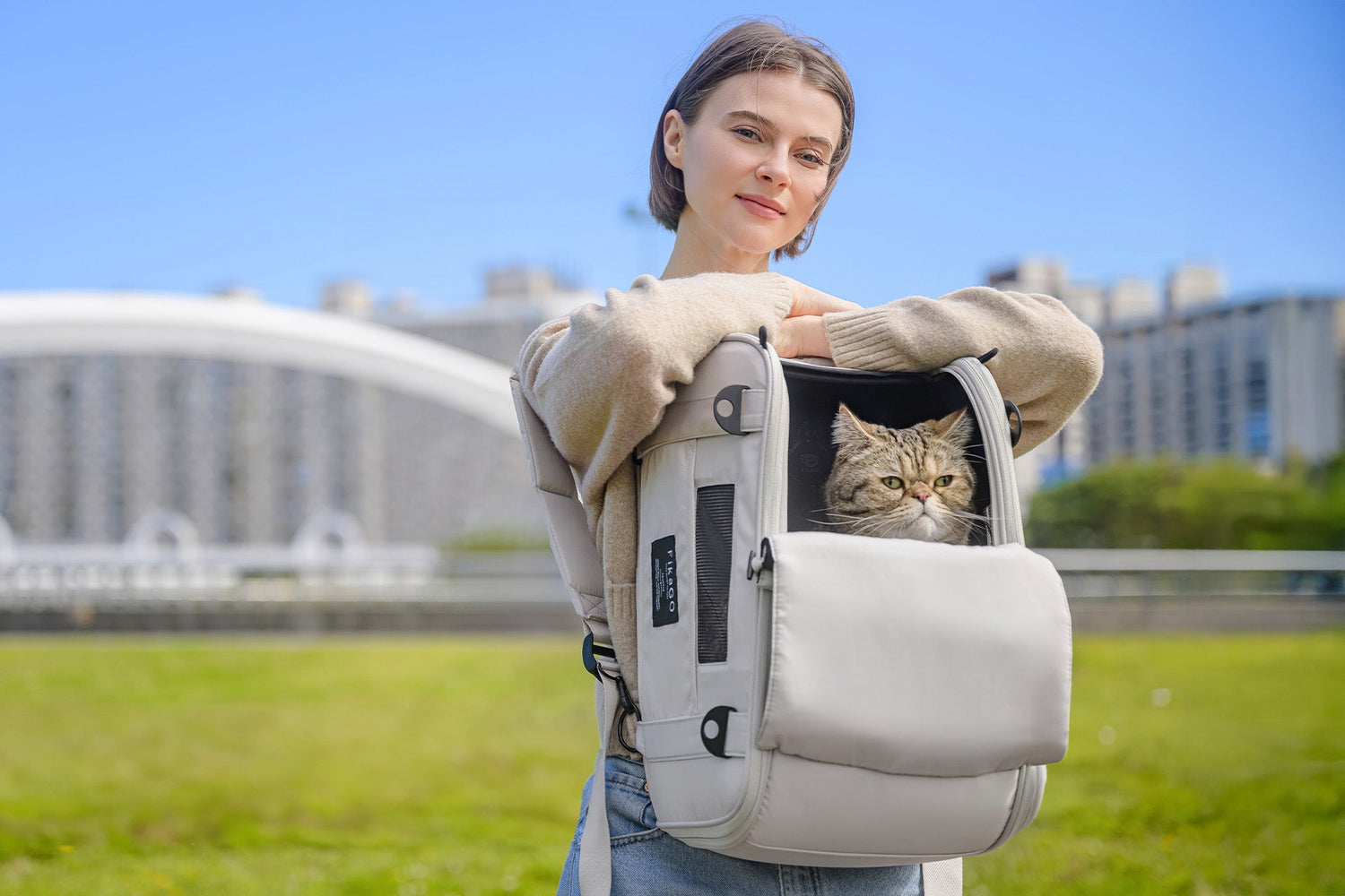 A woman smiles confidently at the camera while wearing a light grey FikaGO pet backpack on her front, with the front flap open to let her tabby cat look out.