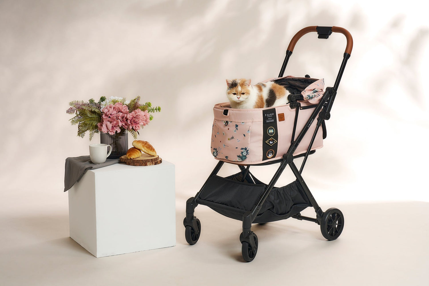A calico cat sitting calmly in a pink FikaGO pet stroller featuring a floral pattern, styled in a studio next to a vase of flowers and pastries.
