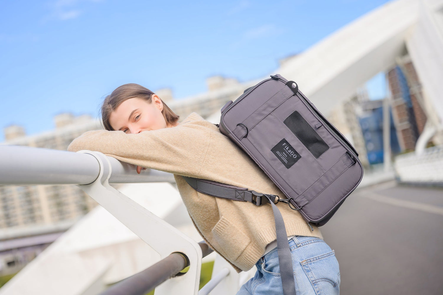 A woman wearing a grey FikaGO pet carrier backpack while standing on a modern city bridge.