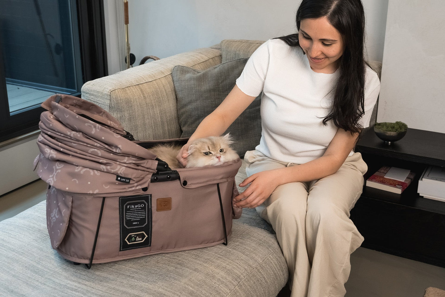 A smiling woman petting her fluffy cat relaxing in a dusty rose FikaGO pet carrier on a sofa.